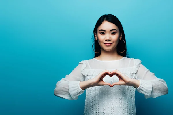 Smiling brunette asian woman showing heart with hands on blue background — Stock Photo