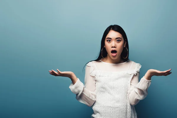 Shocked brunette asian woman showing shrug gesture on blue background — Stock Photo