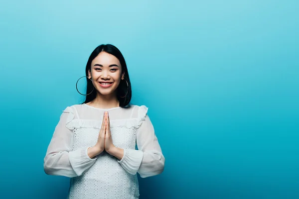 Happy brunette asian woman with praying hands on blue background — Stock Photo