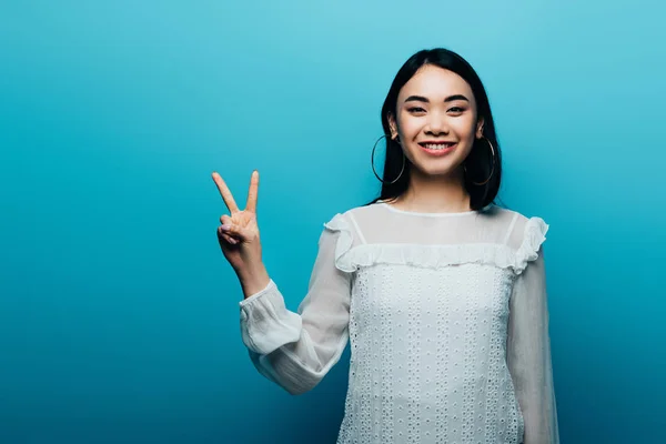 Счастливая брюнетка азиатская женщина показывает знак мира на голубом фоне — стоковое фото