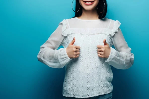 Обрезанный вид счастливой брюнетки азиатской женщины показывая большие пальцы на синем фоне — стоковое фото