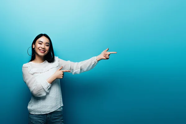 Sorrindo morena asiática mulher apontando com os dedos no fundo azul — Fotografia de Stock