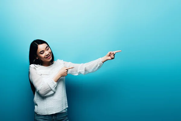 Sorridente bruna donna asiatica che punta con le dita su sfondo blu — Foto stock