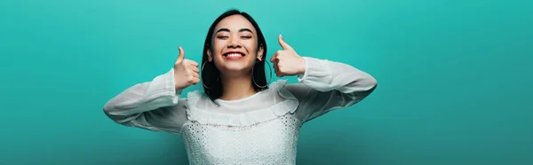 Щаслива брюнетка азіатська жінка, показуючи великі пальці на бірюзовому фоні, панорамний знімок — стокове фото