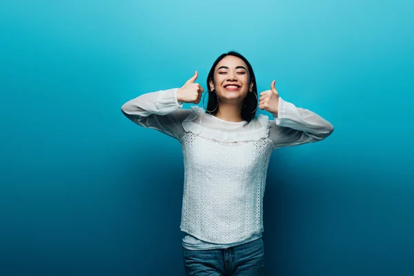 Happy brunette asian woman showing thumbs up on blue background — Stock Photo