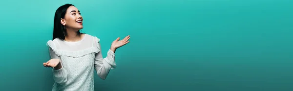 Счастливая брюнетка азиатка, пожимающая плечами на бирюзовом фоне, панорамный снимок — стоковое фото