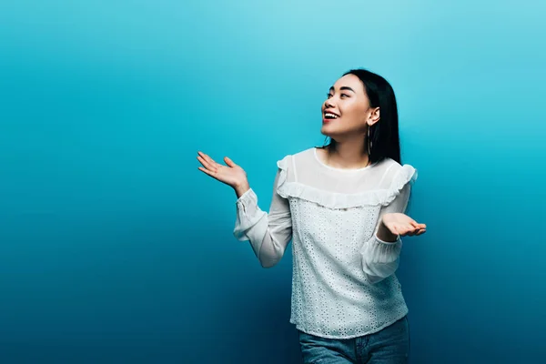 Glücklich Brünette asiatische Frau zeigt Achselzucken Geste auf blauem Hintergrund — Stockfoto