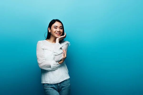 Glücklich Brünette asiatische Frau mit Hand in der Nähe Gesicht auf blauem Hintergrund — Stockfoto