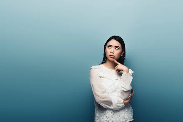 Thoughtful brunette asian woman looking away on blue background — Stock Photo