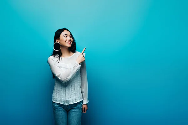 Glücklich Brünette asiatische Frau zeigt mit dem Finger auf blauem Hintergrund — Stockfoto