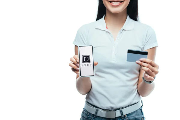 КИЕВ, Украина - 15 июля 2019 года: обрезанный вид улыбающейся брюнетки азиатской девушки с кредитной картой и смартфоном с приложением Uber изолированным на белом — стоковое фото