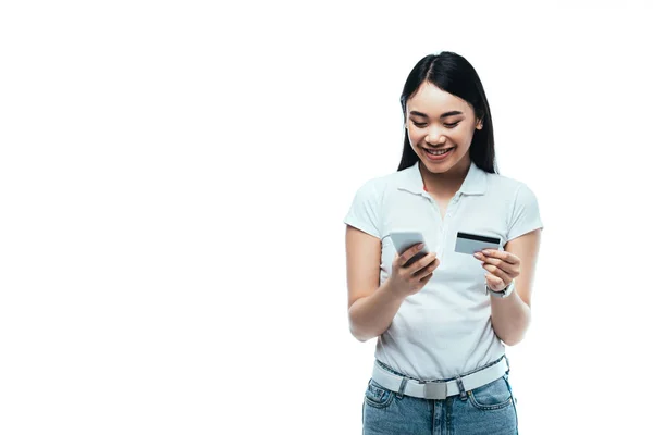 Happy brunette asian girl holding credit card and smartphone isolated on white — Stock Photo
