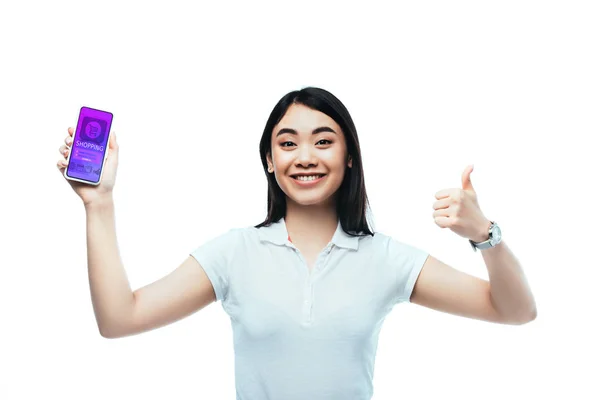 Happy brunette asian woman holding smartphone with online shopping app and showing thumb up sign isolated on white — Stock Photo