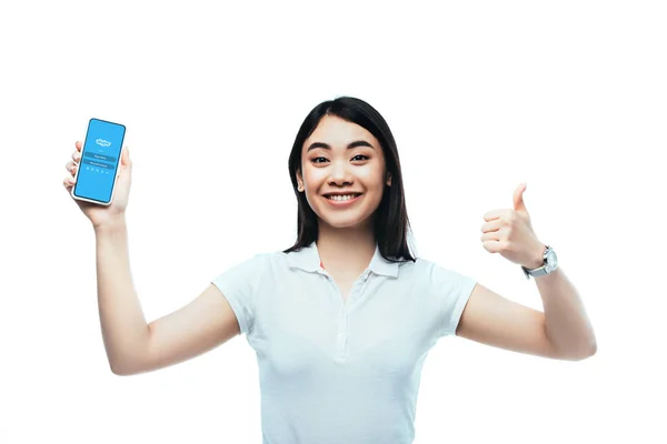 KYIV, UCRAINA - LUGLIO 15, 2019: felice donna asiatica bruna che tiene smartphone con app skype e mostra il segno pollice in alto isolato su bianco — Foto stock
