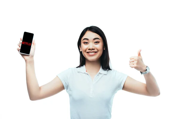 КИЕВ, Украина - 15 июля 2019 года: счастливая брюнетка азиатская женщина держит смартфон с приложением netflix и показывает большой палец вверх знак изолирован на белом — стоковое фото