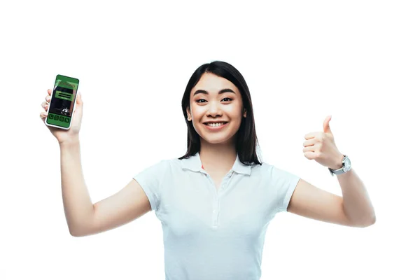 Happy brunette asian woman holding smartphone with booking app and showing thumb up sign isolated on white — Stock Photo