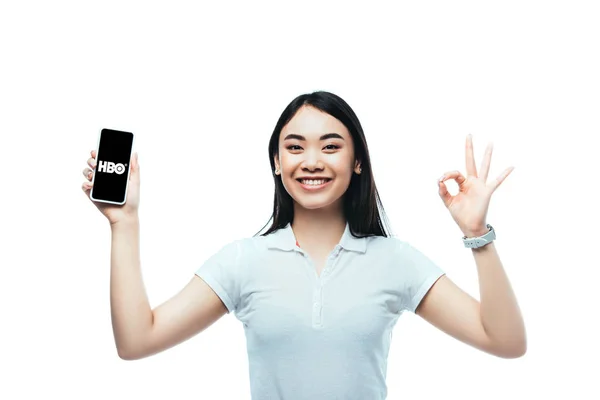 KYIV, UCRANIA - 15 de julio de 2019: morena feliz mujer asiática sosteniendo el teléfono inteligente con la aplicación HBO y mostrando el signo de ok aislado en blanco - foto de stock