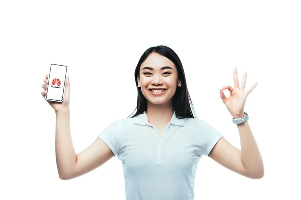 Kyiv, ukraine - 15. Juli 2019: glückliche brünette asiatische Frau hält Smartphone mit Huawei-Logo und zeigt ok Zeichen isoliert auf weiß — Stockfoto