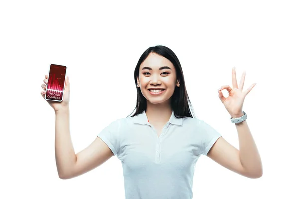 Happy brunette asian woman holding smartphone with trading courses app and showing ok sign isolated on white — Stock Photo