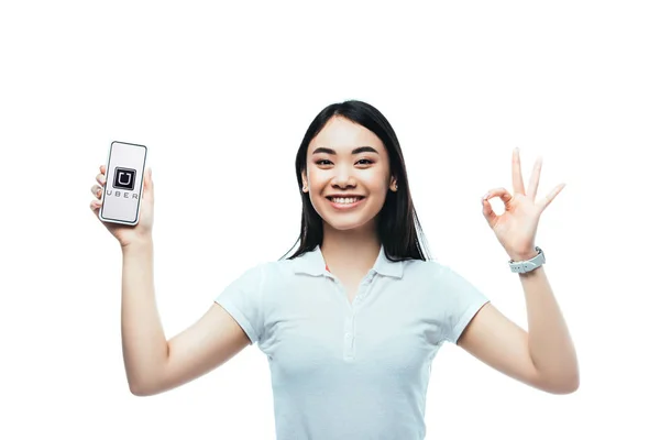 KYIV, UCRANIA - 15 de julio de 2019: morena feliz mujer asiática sosteniendo el teléfono inteligente con la aplicación uber y mostrando el signo ok aislado en blanco - foto de stock