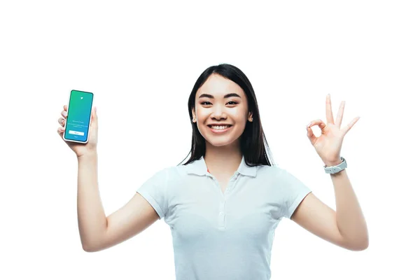 KYIV, UCRAINA - LUGLIO 15, 2019: felice donna asiatica bruna che tiene smartphone con app twitter e mostra il segno ok isolato su bianco — Foto stock