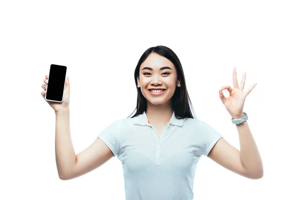Щаслива брюнетка азіатська жінка тримає смартфон з порожнім екраном і показує знак ОК ізольовано на білому — стокове фото