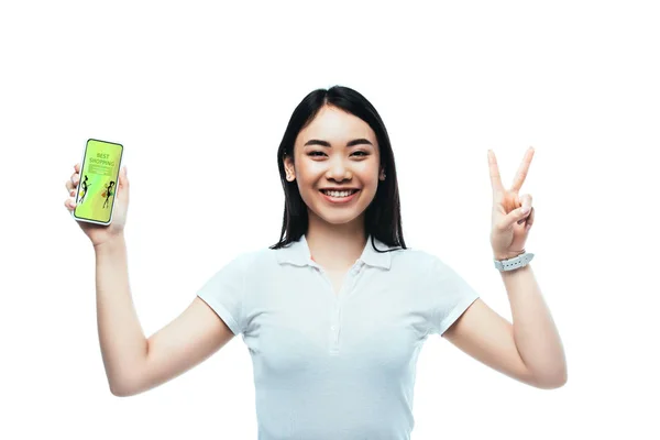 Happy brunette asian woman holding smartphone with best shopping app and showing peace sign isolated on white — Stock Photo