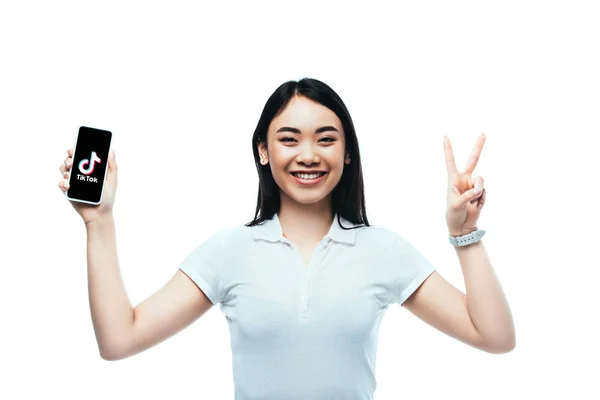KYIV, UCRANIA - 15 de julio de 2019: morena feliz asiática sosteniendo teléfono inteligente con aplicación tiktok y mostrando un signo de paz aislado en blanco - foto de stock