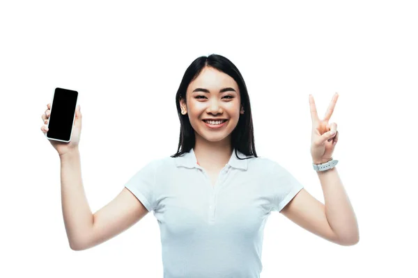 Feliz morena asiática mujer sosteniendo smartphone con pantalla en blanco y mostrando la paz signo aislado en blanco - foto de stock