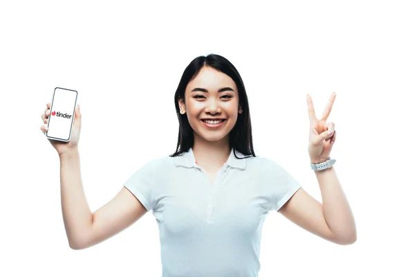 KYIV, UCRAINA - LUGLIO 15, 2019: felice donna asiatica bruna che tiene smartphone con app esca e mostra un segno di pace isolato su bianco — Foto stock