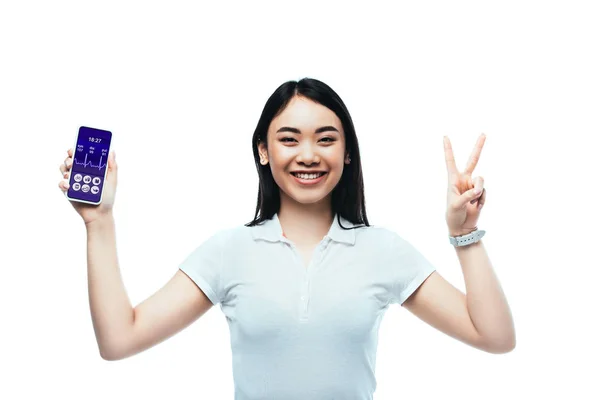 Morena feliz mujer asiática sosteniendo smartphone con aplicación sanitaria y mostrando signo de paz aislado en blanco - foto de stock