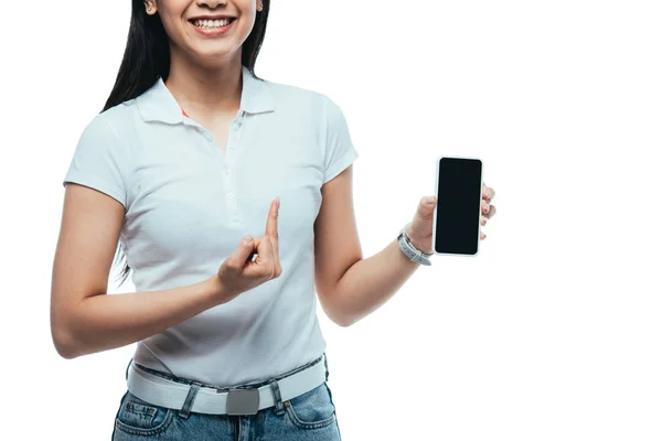 Recortado vista de sonriente morena asiática mujer sosteniendo smartphone con pantalla en blanco y mostrando el dedo medio aislado en blanco - foto de stock
