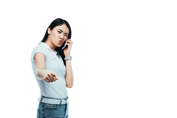 Morena mujer asiática hablando en smartphone y apuntando con el dedo a la cámara aislada en blanco - foto de stock