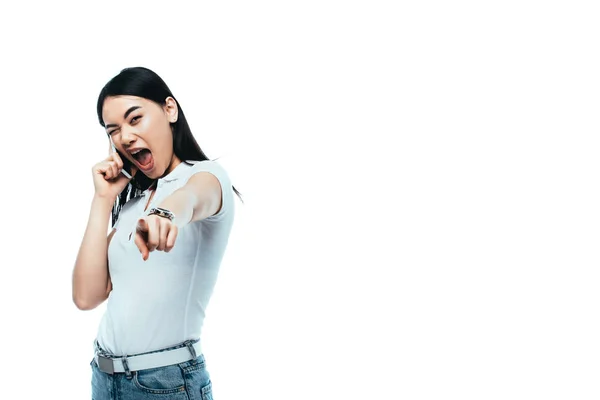 Excited brunette asian woman talking on smartphone and pointing with finger at camera isolated on white — Stock Photo