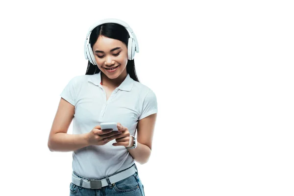 Sorridente attraente ragazza asiatica in cuffia utilizzando smartphone isolato su bianco — Foto stock