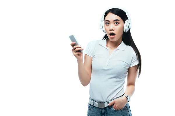 Impactado atractivo asiático chica en auriculares usando smartphone aislado en blanco - foto de stock