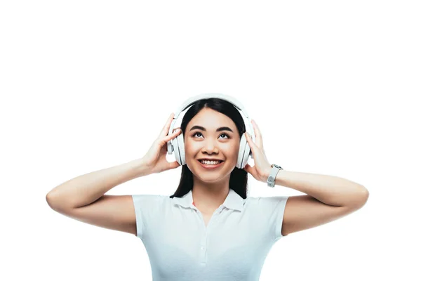Усміхнена приваблива азіатська дівчина в навушниках дивиться вгору ізольовано на білому — стокове фото