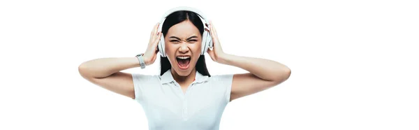 Сердитая привлекательная азиатская девушка кричит в наушниках изолированные на белом, панорамный снимок — стоковое фото