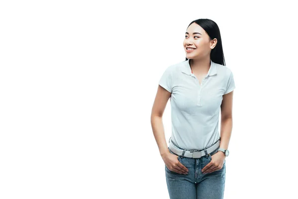 Счастливая привлекательная азиатская девушка с руками в карманах глядя в сторону изолированы на белом — стоковое фото