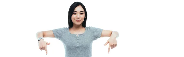 Smiling asian girl pointing with fingers downwards isolated on white, panoramic shot — Stock Photo