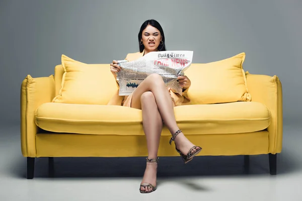 Сердитая азиатская бизнесвумен сидит на желтом диване и читает газету с фейковыми новостями на сером фоне — стоковое фото