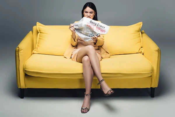 Aggressive asiatische Geschäftsfrau sitzt auf gelbem Sofa und liest Zeitung mit Fake News auf grauem Hintergrund — Stockfoto