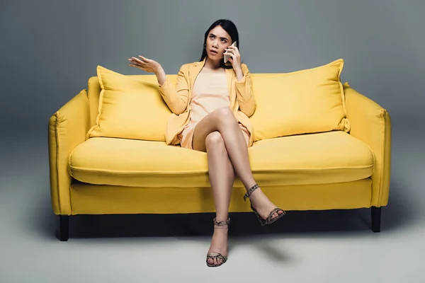 Displeased asian businesswoman sitting on yellow sofa with crossed legs and talking on smartphone on grey background — Stock Photo