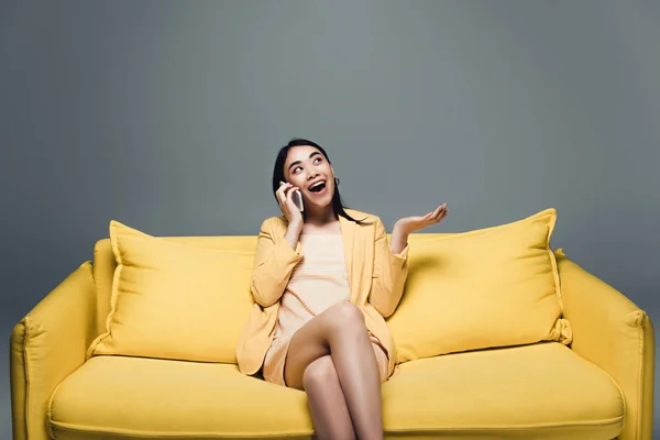 Glückliche asiatische Geschäftsfrau sitzt auf gelbem Sofa mit überkreuzten Beinen und spricht auf Smartphone auf grauem Hintergrund — Stockfoto