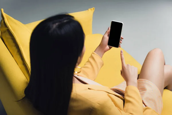 Femme d'affaires brune assise sur un canapé jaune et pointant du doigt un smartphone avec écran blanc isolé sur gris — Photo de stock