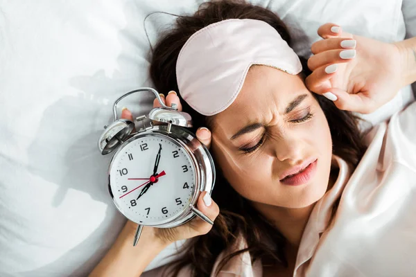 Mujer enojada en la máscara de dormir sosteniendo el despertador por la mañana - foto de stock