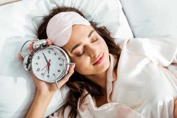 Vista superior de la atractiva mujer con máscara de dormir que sostiene el despertador por la mañana - foto de stock
