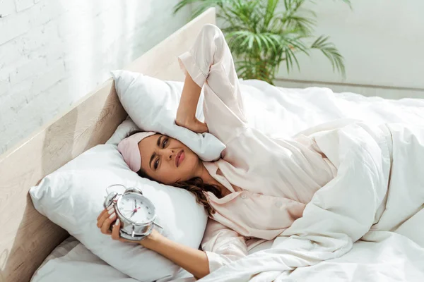 Mujer enojada en la máscara de dormir sosteniendo el despertador por la mañana - foto de stock