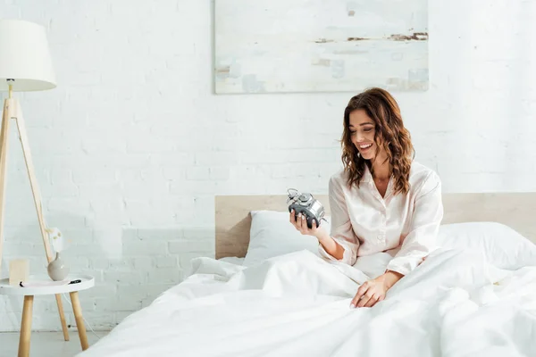 Mulher atraente sorrindo e segurando despertador de manhã — Fotografia de Stock