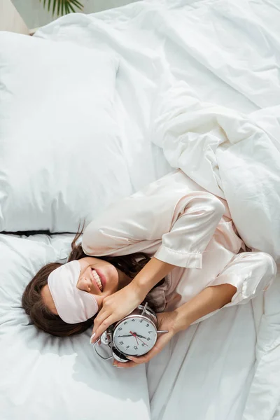 Woman with sleeping mask smiling and holding alarm clock at morning — Stock Photo